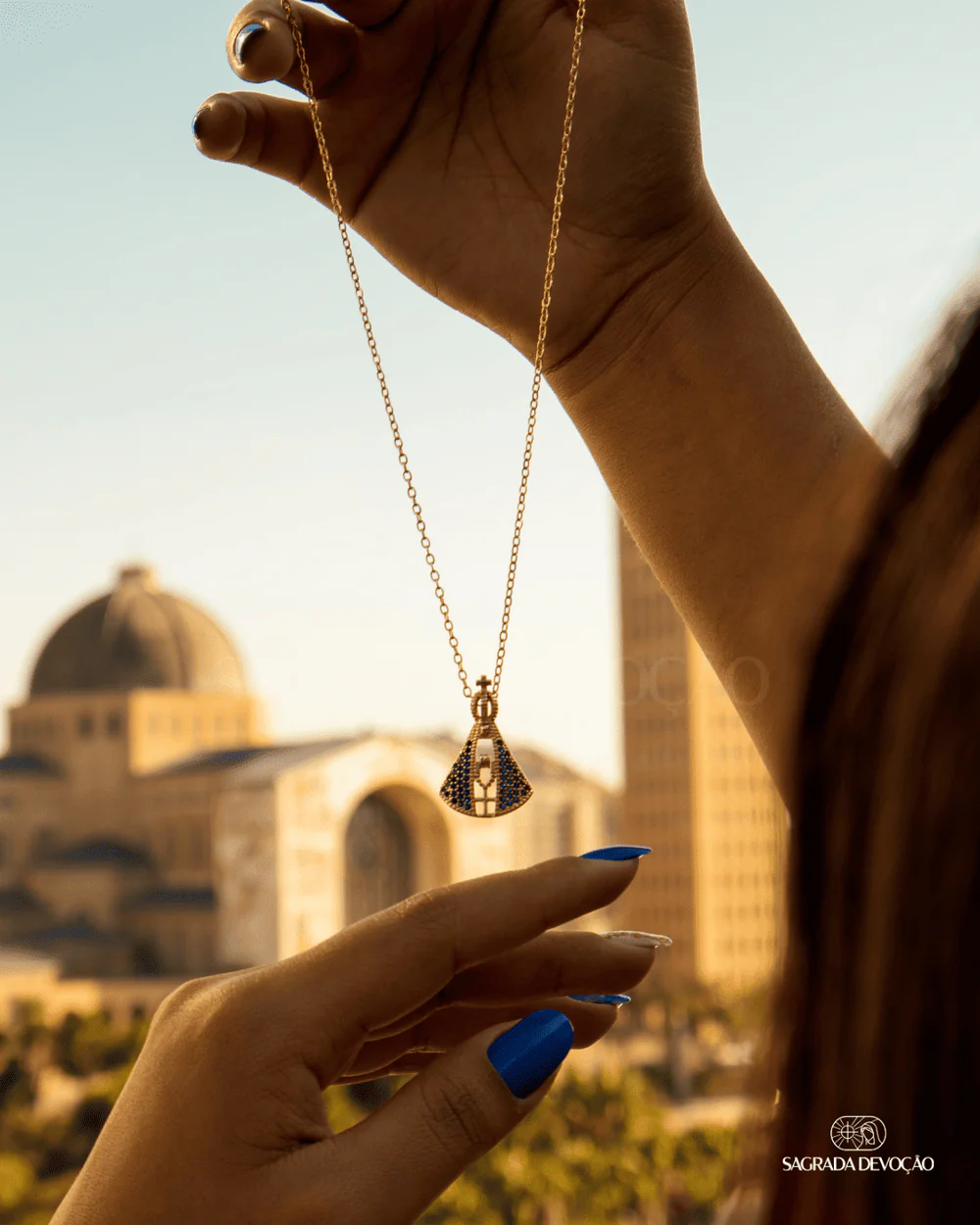 Mãos segurando colar com pingente de Nossa Senhora em frente ao santuário